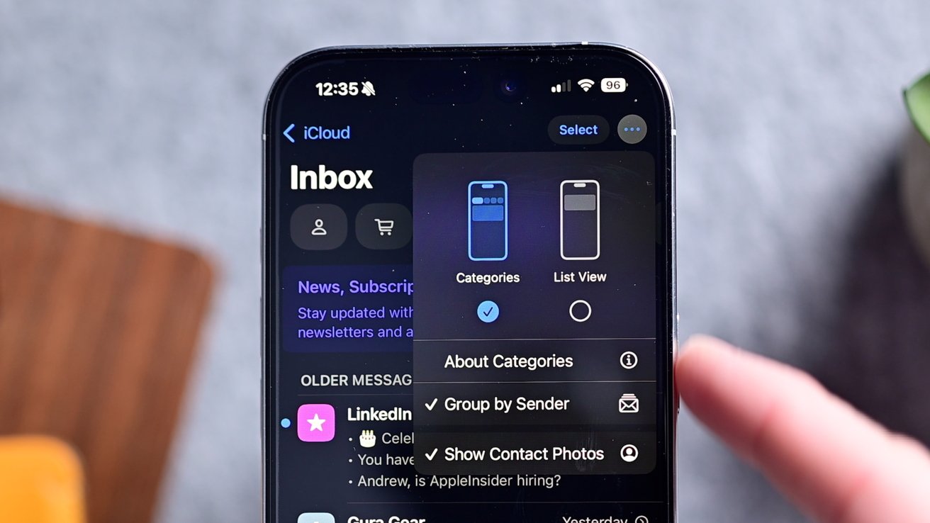 iPhone на сером столе с открытым приложением электронной почты и всплывающим меню с различными опциями, включая переключатель для отображения или скрытия фотографий контактов.